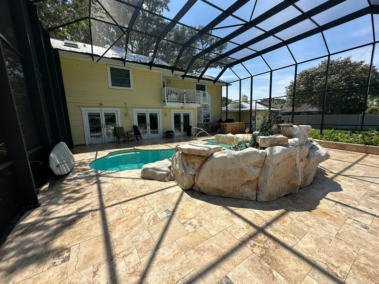 Wide view of a cleaned travertine pool deck with a rock waterfall inside a screen enclosure
